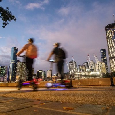 two people on scooters riding along a footpath at dusk. The city skyline is in the background